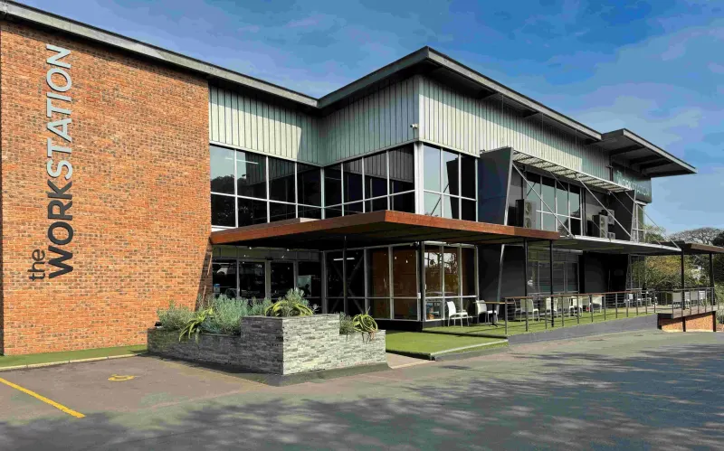 Modern brick and metal building with "the Workstation" signage, glass windows, outdoor seating area, and manicured lawn.