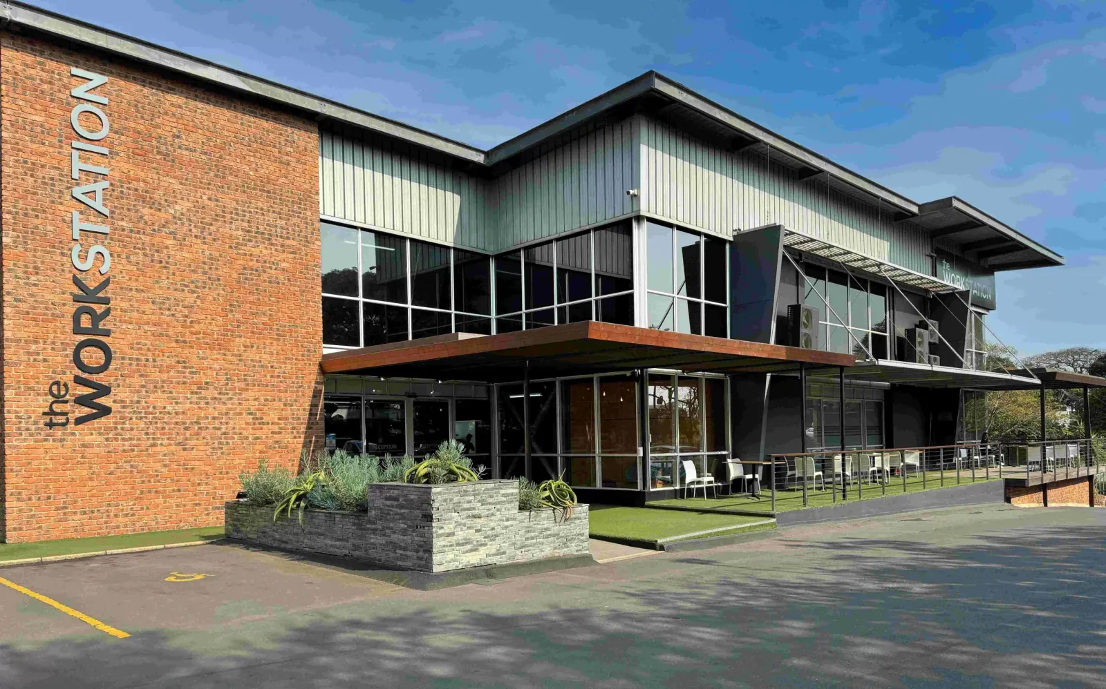 Modern brick and metal building with "the Workstation" signage, glass windows, outdoor seating area, and manicured lawn.
