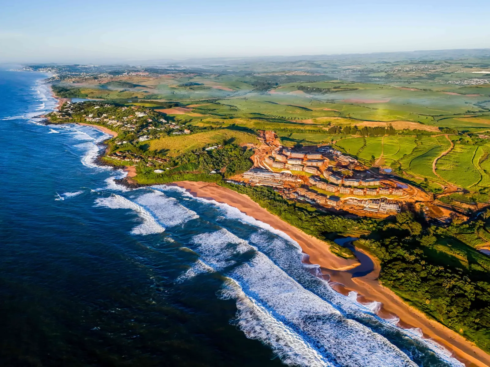 Aerial coastal view showing sandy beach, ocean waves, red cliffs, green farmland, and scattered settlements.